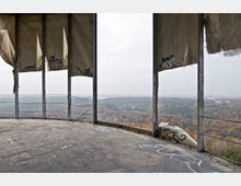 Blick von einer verlassenen Plattform mit Betonboden und beschädigten Stoffwänden auf eine herbstliche Waldlandschaft. Die Metallkonstruktion ist mit Graffiti bedeckt und im Hintergrund ist eine hügelige Landschaft mit einem fernen Fernsehturm zu sehen.