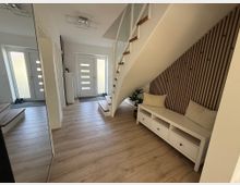 Heller Eingangsbereich eines Hauses mit Holzboden und moderner Treppe. Unter der Treppe befindet sich eine weiße Sitzbank mit Kissen, dahinter eine Wand mit vertikalen Holzpaneelen; im Hintergrund sind eine weiße Eingangstür und seitliche Glasflächen zu sehen.