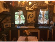 Rustikale Küche mit Holzmöbeln, grünen Fliesen und dekorativen Elementen. Zwei Fenster mit Blumengardinen und Blick ins Grüne, diverse Küchengeräte und eine zentrale Uhr an der Wand, ein Esstisch mit Spitzen-Tischdecke und einem Blumenstrauß im Vordergrund.