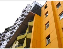 Gelb-orangefarbenes modernes Wohngebäude mit auffälligen Balkonen und geometrischen Fassadenelementen, in kontrastierender Nähe zu einem grauen Gebäudeteil mit dunklen Balkonen.