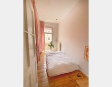 Schmales Schlafzimmer mit hellem Holzboden, weißer Bettwäsche auf einer niedrigen Matratze und einer großen Fensterfront mit Blick auf benachbarte Gebäude. Links hängt ein rosa Vorhang, rechts an der Wand ein gerahmtes Bild, und eine Zimmerpflanze steht im Fensterbereich.