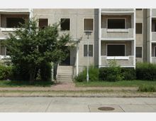 Vorderansicht eines verlassenen Wohngebäudes aus Beton mit leeren Fensteröffnungen. Vor dem Gebäude befindet sich ein Gehweg, eine Straßenlaterne, ein Baum und bewachsene Grünflächen.