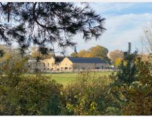 Steinernes Gebäude mit dunkelblauem Dach, umgeben von Wiesen und Bäumen, im Vordergrund rahmen Zweige und Büsche die Szenerie ein. Der Himmel ist klar, und die Bäume zeigen Herbstfarben.