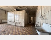 Verlassener Sanitärraum mit weiß gefliesten Wänden und Trennwänden, freiliegenden Rohrleitungen sowie einer alten Badewanne im Vordergrund. Der Boden ist kariert gefliest, und die Decke zeigt großflächig abblätternden Putz.