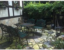 Kleiner Gartenhof mit einer Terrasse aus unregelmäßigen Steinplatten, ausgestattet mit grünen Metallstühlen und einer Bank um einen Tisch. Im Hintergrund wachsen dichte Weinranken auf einer Pergola, daneben stehen Topfpflanzen und eine Laterne zur Dekoration.