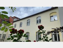 Gelb gestrichene Fassade eines mehrstöckigen Gebäudes mit weißen Fensterrahmen und Rundbogenfenstern, umgeben von roten Rosen und grünem Laub im Vordergrund. Der Himmel ist bewölkt.