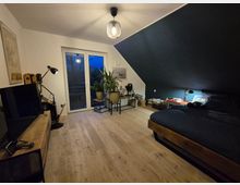 Schlafzimmer mit dunkler Akzentwand, Holzboden und minimalistischer Einrichtung. Ein Bett mit dunkler Bettwäsche steht rechts, links ein Fernseher auf einem Holztisch, daneben mehrere Pflanzen vor einer Balkontür.
