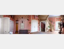 Historischer Eingangsbereich mit rosa Wänden, dekoriert mit zahlreichen Jagdtrophäen und Geweihen an den Wänden. Der Raum enthält eine hölzerne Treppe, eine hohe Standuhr, weiße Doppeltüren und antike Möbel, darunter eine Truhe und ein kleiner Tisch.