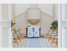 Innenansicht einer kleinen, hellen Kapelle mit einem Altarbereich. Der Raum ist mit Holzstühlen und weißen Dekorationen für eine Hochzeit hergerichtet; im Hintergrund befindet sich eine dekorative, beige Kanzel vor einer schlichten Wand.