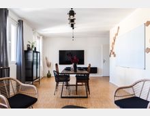 Wohnzimmer oder Besprechungsraum mit hellem Holzboden, einem Esstisch mit Stühlen in der Mitte, einer schwarzen Hängelampe, einem großen wandmontierten Fernseher und einem Regal mit Glasfront. An der Wand befinden sich eine weiße Tafel und geometrische Holzdekorationen.