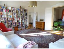 Wohnzimmer mit Bücherregal an der Wand, einem roten Sessel, einer Gitarre, und einem weißen Sofa. In der Mitte liegt ein großer, gemusterter Teppich, daneben eine Holzkommode und ein Topfpflanze. Eine breite Tür führt zu einem angrenzenden Raum mit Fenstern und Vorhängen.