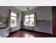 Ein leerer Raum mit zwei großen Fenstern, die mit beigen Vorhängen versehen sind, und darunter angebrachten Heizkörpern. Links ist ein kleines Waschbecken mit einem Spiegel sichtbar, während der Boden aus glänzendem, dunkelrotem Material besteht.