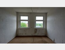 Leerer Raum mit zwei großen Fenstern, durch die Bäume und Teile eines Gebäudes sichtbar sind. Die Wände sind schlicht und hell, zeigen jedoch Schmutzspuren, während der Boden abgenutzt wirkt.