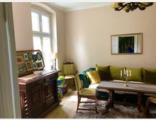 Wohnzimmer mit klassischer Einrichtung: Ein hölzernes Sideboard mit Bilderrahmen und einer Lampe steht vor einem hohen Fenster. Ein grünes Sofa mit farbigen Kissen und ein Holztisch mit Kerzenständer befinden sich auf einem Teppich. Ein Spiegel hängt an der Wand.