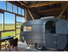 Ein kleiner, grauer Wohnwagen steht in einem offenen, rustikalen Raum mit Holzbalken und großen Fenstern, die den Blick auf eine grüne Wiese und blauen Himmel bieten. Daneben stehen ein Holztisch und ein Stuhl auf einem Teppich.