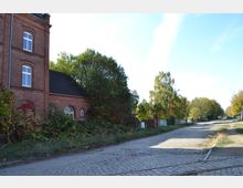 Ehemaliges Backsteingebäude mit weiß-gerahmten Fenstern und einer Fassade, die mit Kletterpflanzen bewachsen ist, neben einer gepflasterten Straße in einer baumreichen, ländlichen Umgebung.