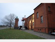 Backsteingebäude mit hohen Fenstern und gepflastertem Weg, daneben ein freistehender Wasserturm aus rotem Backstein mit grünem Dach. Im Vordergrund steht ein Hebebühnenfahrzeug mit Beleuchtungsausrüstung, daneben eine Grünfläche mit kahlem Baum.