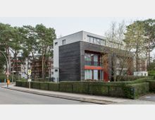 Moderne Wohngebäude mit Holz- und Putzfassade, großen Fenstern und Balkonen, umgeben von Bäumen und Hecken. Im Vordergrund eine Straße mit Bürgersteig, Verkehrsschildern und Bushaltestelle.