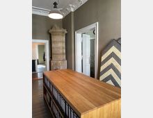 Innenraum eines historischen Büros mit hohen Decken und dekorativer Stuckverzierung. Im Vordergrund steht ein großer Holztisch mit Aktenordnern darunter, im Hintergrund ein kunstvoll verzierter Kachelofen sowie offene weiße Doppeltüren, die in einen weiteren Raum führen.