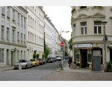 Straßenszene in einer Stadt mit historischen Gebäuden, einem Café namens "Sophieneck" an der Ecke und parkenden Autos entlang der Straße. Die Fassade der Gebäude ist beige und hellgrau, mit hohen Fenstern und dekorativen Details, im Hintergrund ist ein Baustellenkran sichtbar.