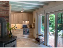 Küche mit hellen Holzschränken, moderner Einbauküche und Keramikfliesenboden, neben einer großen Fensterfront mit Blick auf eine Terrasse mit Gartenmöbeln und viel Grün. Links im Vordergrund ein Kaminofen aus Metall und eine Backsteinwand.