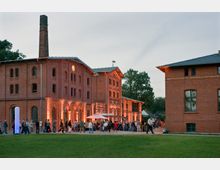 Ziegelrotes, historisches Industriebauwerk mit mehreren Etagen, großen Fenstern und einem hohen Schornstein, beleuchtet durch warmes Licht; im Vordergrund Menschen, die sich vor dem Gebäude aufhalten, sowie eine Wiese.