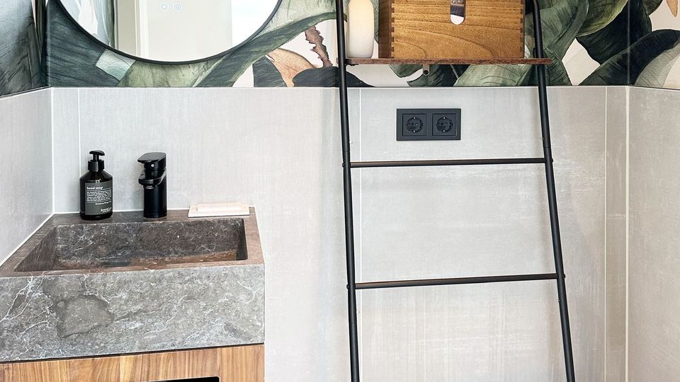 Modernes Badezimmer mit einer grauen Steinwaschbeckenplatte, einem Unterschrank aus Holz, einer schwarzmetallenen Leiter als Handtuchhalter, und tropisch bedruckter Tapete mit großen Blättern. Im Hintergrund ist eine helle Wand mit Steckdosen sichtbar.