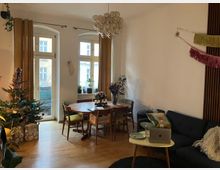 Wohnzimmer mit Holzboden, einer Sitzecke mit dunklem Sofa und einem kleinen Couchtisch sowie einem Esstisch mit vier Stühlen. Große Fenster mit gelben Vorhängen lassen Tageslicht herein, und ein geschmückter Weihnachtsbaum steht links neben der Balkontür.