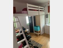 Ein Kinderzimmer mit einem Hochbett aus weißem Holz, einer Leiter und einem Schreibtisch darunter. Der Raum hat helle Holzböden, bunte Vorhänge und diverse Möbel wie eine Kommode und einen Teppich.