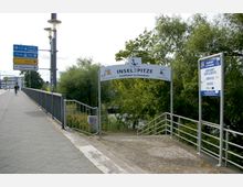 Eine Gehwegbrücke mit Geländer führt zu einer Beschilderung, die den Eingang der Schiffsanlegestelle „Inselspitze – Schifffahrt in Potsdam“ markiert. Im Hintergrund sind Bäume und Wegweiser zu verschiedenen Städten zu sehen.