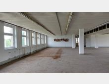 Leerer Innenraum eines verfallenen Gebäudes mit weißen Wänden, mehreren rechteckigen Fenstern auf der linken Seite, einer abgenutzten Betonbodenfläche und sichtbaren Rohren unterhalb der Fenster. Die Decke ist mit Metallpanelen verkleidet, und einzelne Säulen stützen den Raum.