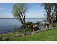 Eine Uferlandschaft mit Blick auf einen großen See, umgeben von Bäumen und Wiesen. Im Vordergrund stehen eine Sitzgruppe aus Holzstühlen und ein Tisch, teils mit einer Abdeckung, auf einer Rasenfläche mit Steinbegrenzung.