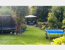 Ein gepflegter Garten mit einer Rasenfläche, einer Tischtennisplatte, einem Trampolin und einem Baum mit Kletterpflanzen. Im Hintergrund befinden sich Sitzmöbel auf einer Terrasse mit Sonnenschirm sowie ein Essbereich, umgeben von dichten Büschen und Blumenbeeten.