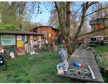 Ein alternativer Gartenbereich mit Holzbauten, Schildern und Dekorationen, umgeben von Bäumen und Wiese. Im Vordergrund befinden sich ein langer Holztisch mit Seilen und einer Büste sowie ein Fahrrad.