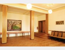 Ein heller Raum mit Parkettboden, Holzwandverkleidungen und einer kleinen erhöhten Bühne mit Treppenstufen. An den Wänden hängen zwei Kunstwerke, darunter ein großes Bild mit einem Baummotiv, und auf der rechten Seite befinden sich gepolsterte Stühle.