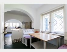 Helle Küche mit Eckbank und Holztisch vor einer Fensterfront mit Sprossenfenstern; im Hintergrund ein Durchgang mit Rundbogen führt in den Bereich mit Kochinsel, Schachbrettmuster-Fliesenboden und Arbeitsflächen.