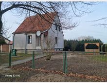 Ein Einfamilienhaus mit hellem Putz und rotem Satteldach, umgeben von einem Garten mit kahlen Bäumen. Im Vordergrund ein Metallzaun, daneben ein Holzpavillon und eine Hecke im Hintergrund.