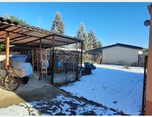 Schneebedeckter Garten mit einem Carport auf der linken Seite, unter dem ein Fahrrad und eine abgedeckte Ladung stehen. Im Hintergrund eine weiße Backsteinmauer, Nadelbäume und ein flaches Gebäude.