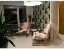 Ein gemütlich eingerichteter Wohnbereich mit zwei Schaukelstühlen aus Holz und Rattan, die mit weißen Fellauflagen dekoriert sind. Im Hintergrund befinden sich eine Glas-Schiebetür, eine Wand mit tropisch bedruckter Tapete sowie eine Zimmerpflanze und ein Beistelltisch mit Dekoration.