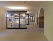 Innenraum eines Museums mit beleuchteten Vitrinen, in denen architektonische Modelle, Texttafeln und Exponate ausgestellt sind. Rechts befinden sich weiße Rundbögen mit rotbraunen Steinpfeilern, während der Boden aus beigen Fliesen besteht.