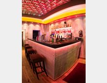 Moderne Bar mit Theke aus rustikalem, hellgrauem Holz und schwarzer Arbeitsfläche, hinter der diverse Flaschen und Gläser auf Regalen beleuchtet sind. Die Decke ist mit einem dekorativen Muster aus roten und gelben Lichtern gestaltet, und rote Hocker stehen vor der Theke auf einem Holzboden.