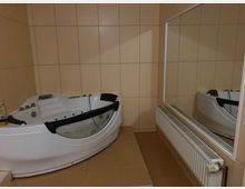 Eckbadewanne in einem Badezimmer mit beigen Fliesen, großem rechteckigem Wandspiegel und darunterliegendem Heizkörper. Die Badewanne verfügt über schwarze Armlehnen und eingelassene Armaturen.