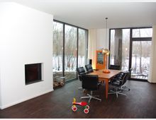 Esszimmer mit Holztisch und sechs schwarzen Ledersesseln, großen bodentiefen Fenstern, die einen Blick auf einen verschneiten Außenbereich bieten, und einer weißen Wand mit eingebautem Kamin. Auf dem dunklen Holzfußboden steht ein rotes Kinderrad.