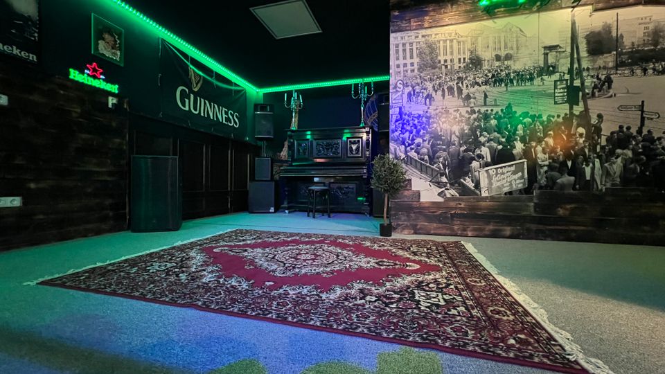 Innenraum einer Bar oder eines Veranstaltungsraums mit dunklen Holzpaneelen, grünem LED-Deckenlicht und Leuchtschildern. Im Hintergrund stehen ein Klavier mit Lautsprechern, an der Wand hängt ein großes Schwarzweiß-Fotomural, und auf dem Boden liegt ein großer gemusterter Teppich.