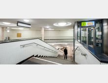 U-Bahn-Station mit einer Treppe, die zu einem unteren Bahnsteig führt, einer Aufzugstür mit der Beschriftung "U9" rechts und hellen, deckenmontierten Lampen. Die Wände und das Geländer der Treppe sind aus weißen Fliesen mit Metallhandläufen.