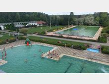 Ein Freibad mit mehreren Schwimmbecken, darunter ein großes Becken im Vordergrund mit Badegästen und einer kleinen grünen Rutsche. Im Hintergrund befinden sich ein weiteres langes Schwimmbecken, Liegeflächen mit Bänken, eine Rasenfläche, Bäume und Gebäude mit rotem Dach.