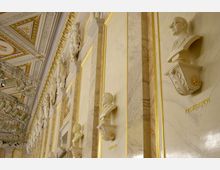 Innenraum einer prunkvollen Halle mit Wandverkleidungen aus hellem Marmor, goldenen Verzierungen und Büsten bekannter Persönlichkeiten in regelmäßigen Abständen. Die Decke ist ornamental gestaltet und mit goldenen Details sowie Fresken geschmückt.