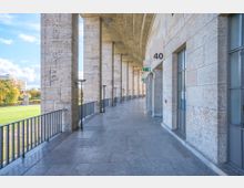 Steinerne Arkadengänge mit hohen rechteckigen Säulen und einem gewölbten Dach, die entlang einer modernen Fassade verlaufen. Auf der linken Seite befinden sich ein Geländer und ein Blick auf eine grüne Rasenfläche mit einigen Bäumen und einem Kunstwerk.