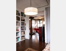 Heller Wohnraum mit offener Gestaltung, bestehend aus einem Bereich mit eingebauten Bücherregalen, Holzboden und einer großen Hängelampe. Im Hintergrund ist ein gemütlicher Sitzbereich mit roten Sofas und einem Holzbalken zu sehen, umgeben von freiliegenden Backsteinwänden und großen Fenstern, die viel Tageslicht hereinlassen.