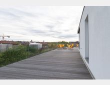 Rooftop-Terrasse mit Holzboden, gelben Liegestühlen und einem transparenten Glasgeländer, umgeben von grünen Baumkronen mit Blick auf eine städtische Skyline im Hintergrund. Eine weiße, moderne Gebäudewand befindet sich rechts im Bild.